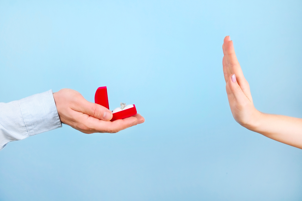 A hand holding an engagement ring in a box and another hand being held up to it to say 'no'